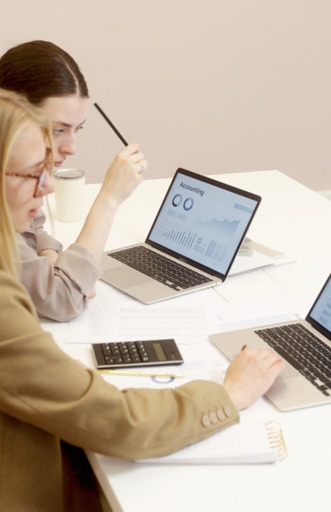 Two women collaborate on financial data analysis with laptops in an office setting.