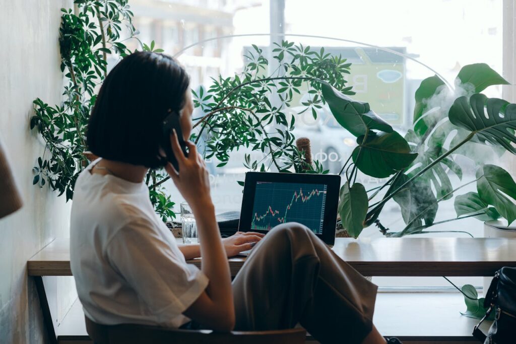 Businesswoman on phone, analyzing stock graphs on laptop in a café.